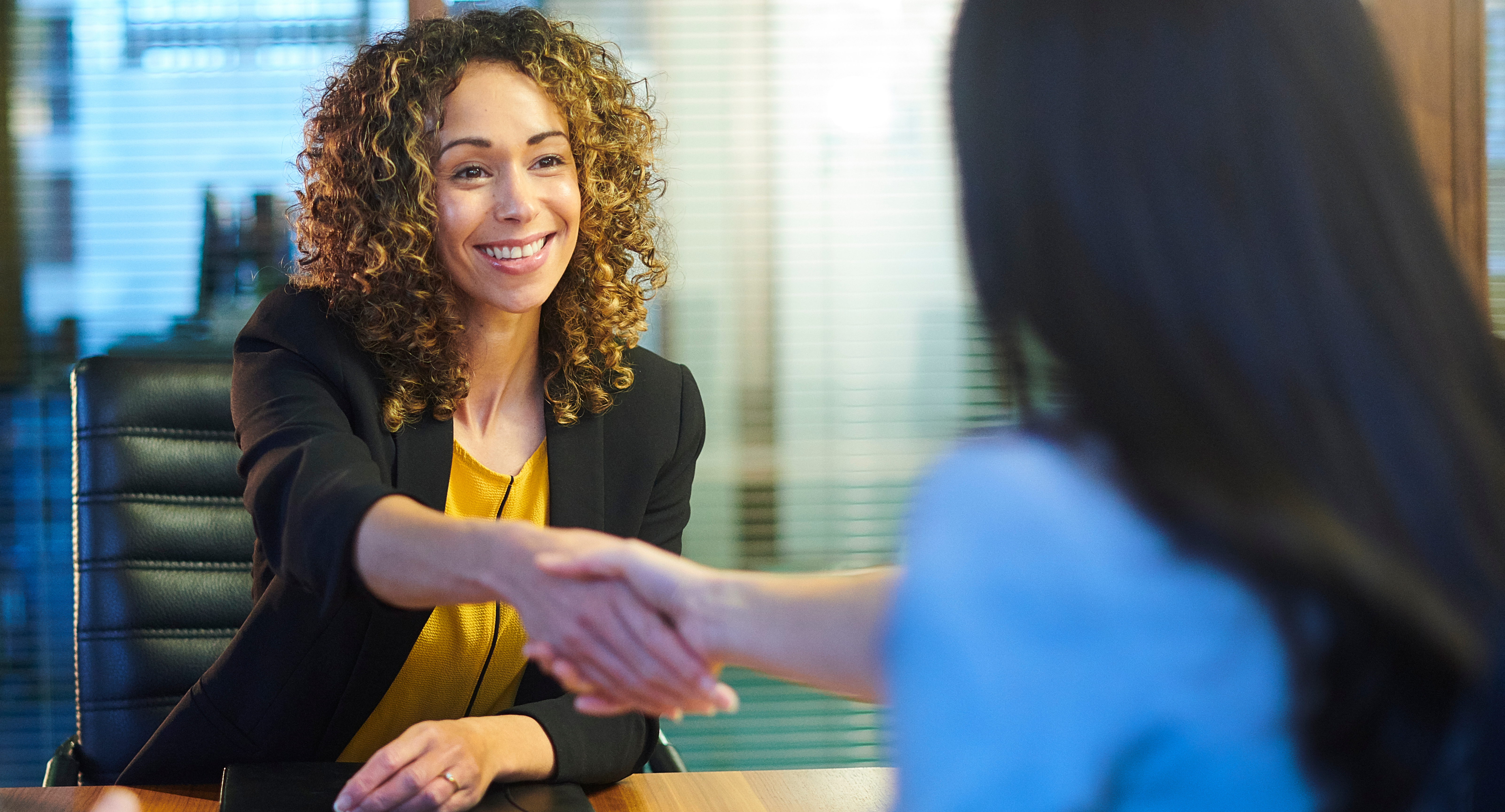 A woman with curly hair smiling while shaking the hand of another woman at the start of an interview.