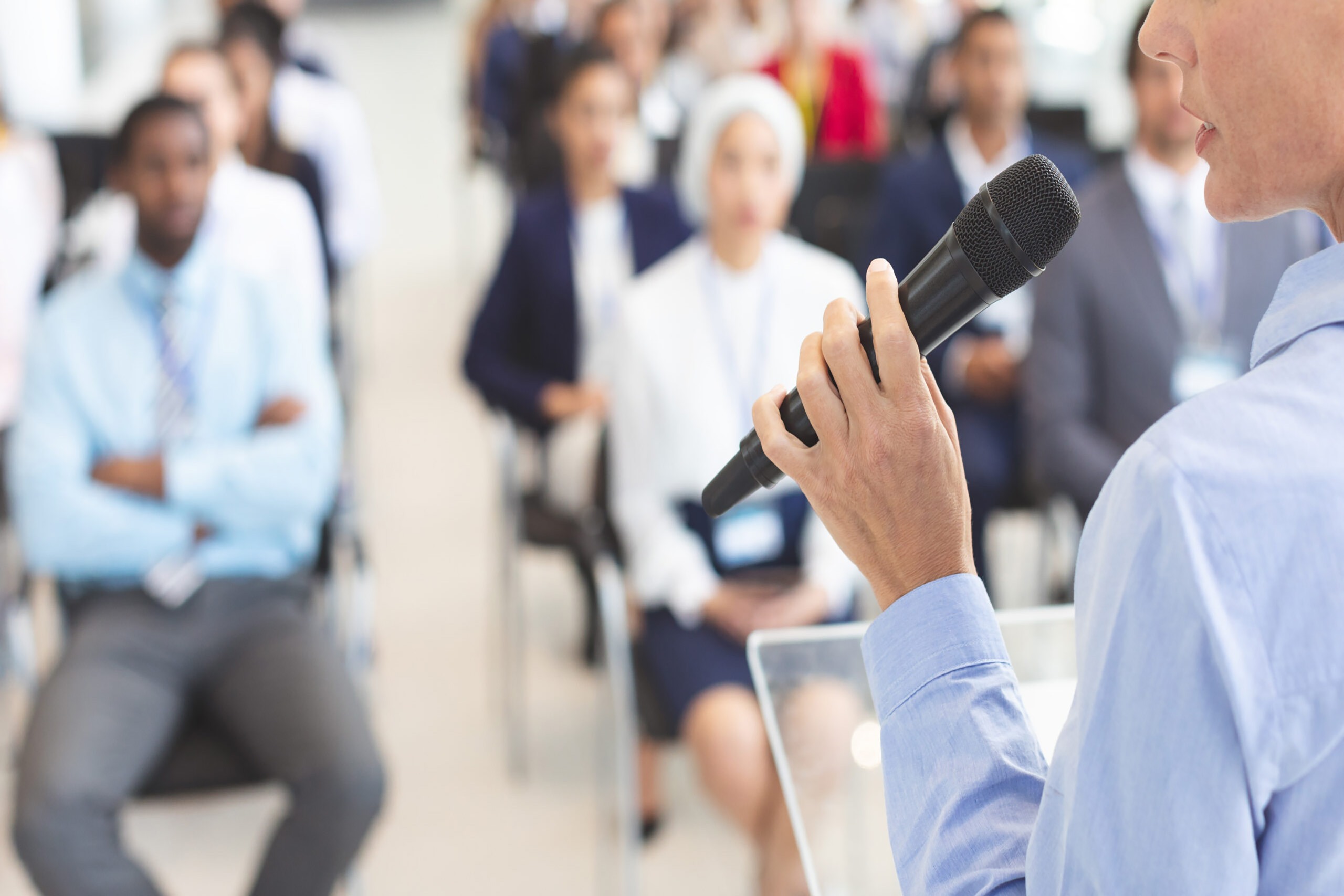 Partial view of a woman holding a microphone while speaking to a diverse audience.